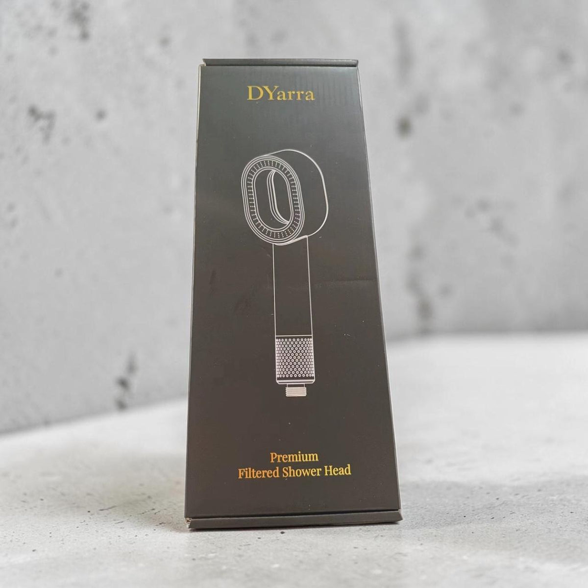 A tall black product box for DYarra Premium Filtered Shower Head, featuring a white line drawing of the shower head and gold text, standing on a light gray surface against a textured gray wall.